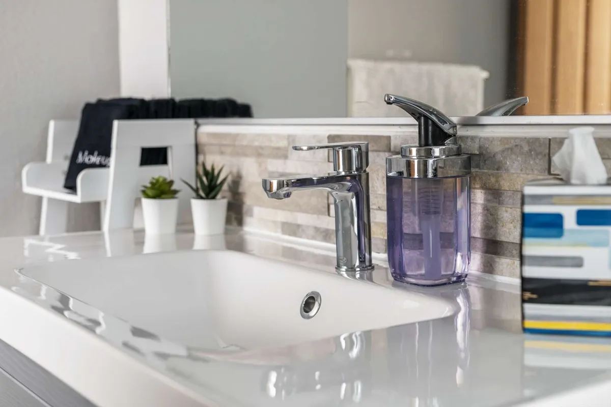 The image captures a refreshing and serene interior view of a bathroom sink area, featuring a modern faucet, a soap dispenser, green plants, and a tissue box. Natural light bathes the scene, highlighting a peaceful space perfect for self-care routines.