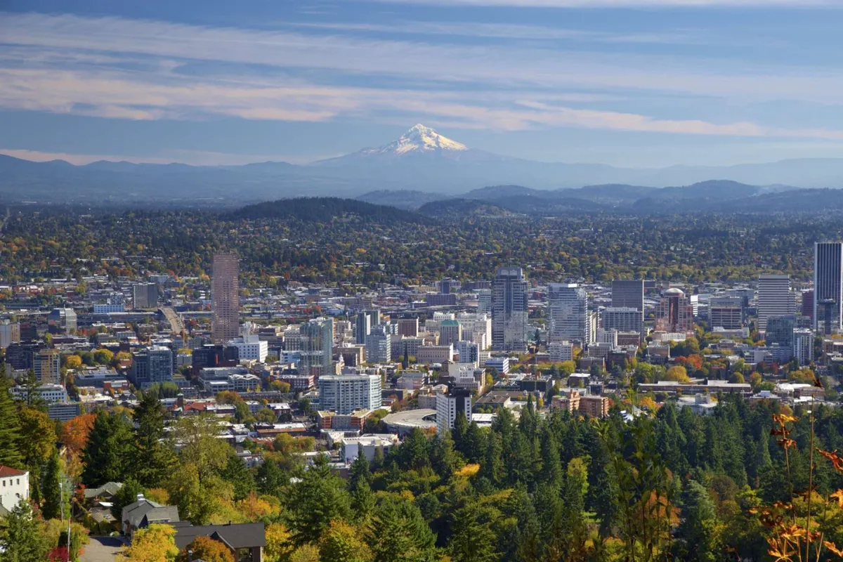 A sweeping view of the Portland city skyline with Mount Hood in the distance, surrounded by lush green foliage and bursts of fall color. The scene evokes a sense of adventure and connection with both urban and natural elements, suggesting easy access to outdoor exploration and breathtaking vistas.