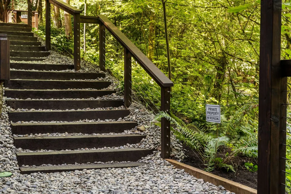 This image captures exterior wooden stairs surrounded by lush greenery, with a private property sign indicating exclusivity. The scene is tranquil, showing the descent from the entryway into a peaceful, forested environment. The dense foliage and natural setting evoke the sensation of arriving at the edge of the woods, perfectly matching the narrative of a forest-edge retreat.