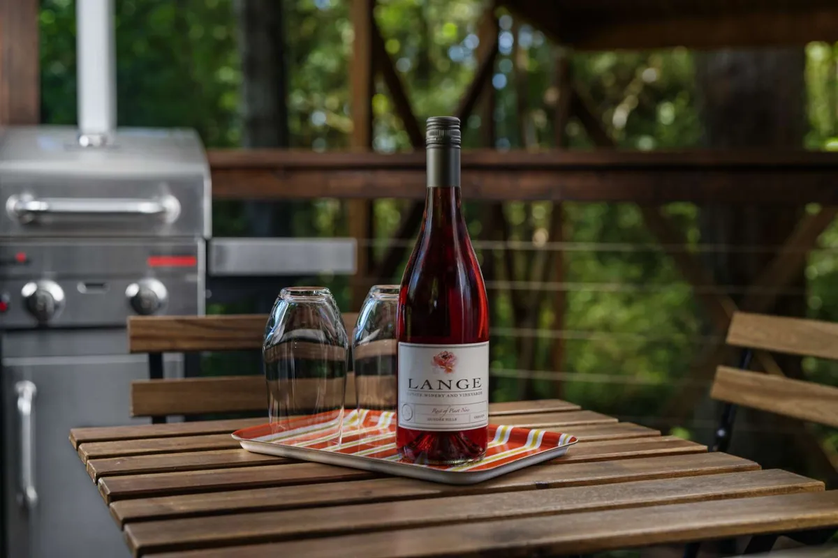 This image shows an outdoor table set with a wine bottle and glasses, accompanied by a grill, suggesting a space for socializing and al fresco dining. The casual setup underlines the potential for memorable evenings on the deck, nestled in the forest setting.