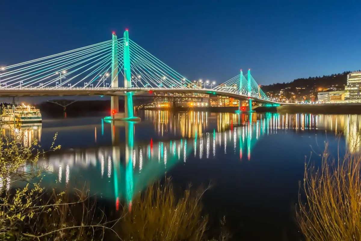 A tranquil evening scene featuring an illuminated bridge, shimmering reflections on the water, and glowing city lights. The calm and picturesque mood highlights the allure of exploring Portland's outdoor spaces after dark, with opportunities for photography and gentle strolls along well-lit paths.