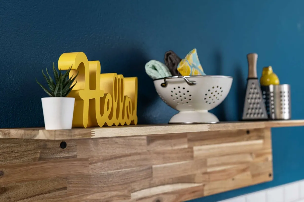 A bright, cheerful kitchen with a welcoming 'Hello' sign, a white colander filled with kitchen utensils, and a small potted plant. The scene exudes warmth and sociability, suggesting a space where friends and family can gather to cook or share a meal.