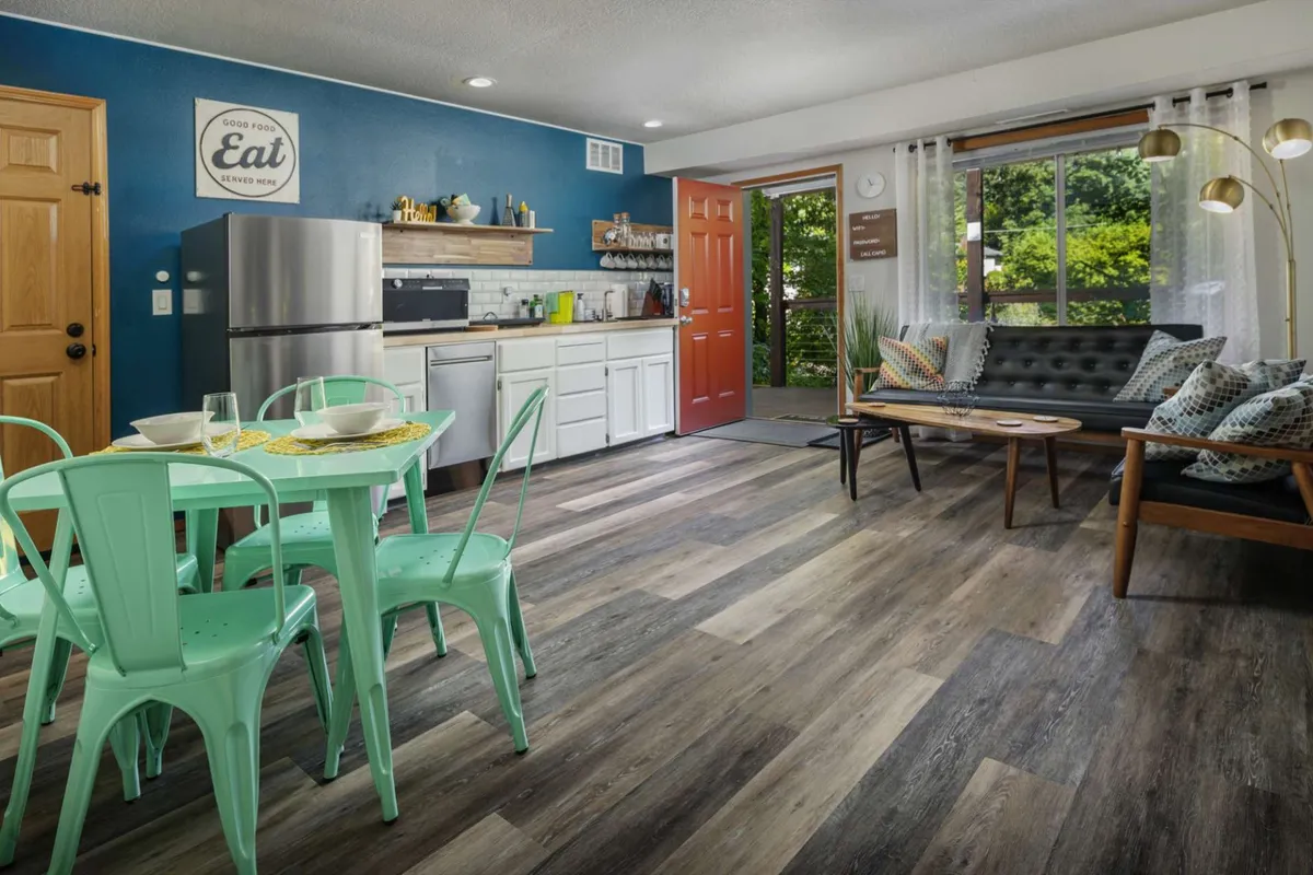The image features a stylish, welcoming dining and living area with vibrant green chairs around a wooden table, a comfortable black sofa, and a striking blue accent wall. Natural light filters in, creating a cozy space perfect for intimate meals, relaxing with a book, or unwinding with family after a day exploring Portland or the nearby parks.