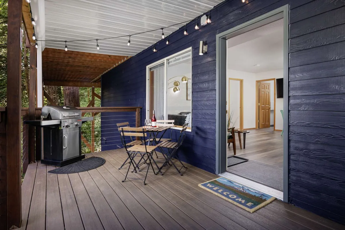 This image features a covered deck with a bistro table, grill, string lights, and a welcome mat. The seating area looks inviting and is suggestive of both morning relaxation and evening gatherings, with forest views implied by the outdoor setting. The overall mood is welcoming and cozy, perfect for savoring coffee or unwinding with wine as mentioned in the content.