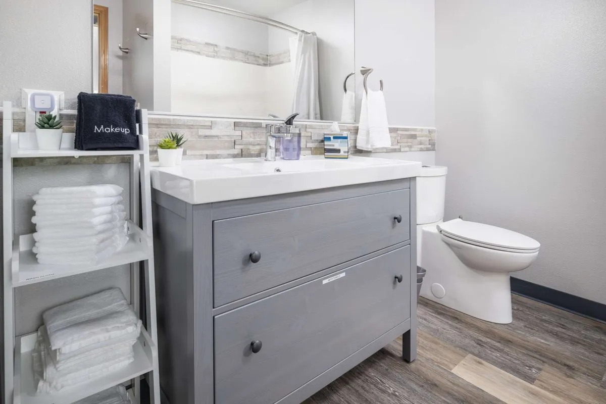 This image focuses on a modern full bathroom with a sleek vanity, large mirror, toilet, and a neatly organized shelving unit. The space feels refreshing and spa-like, ideal for self-care routines.